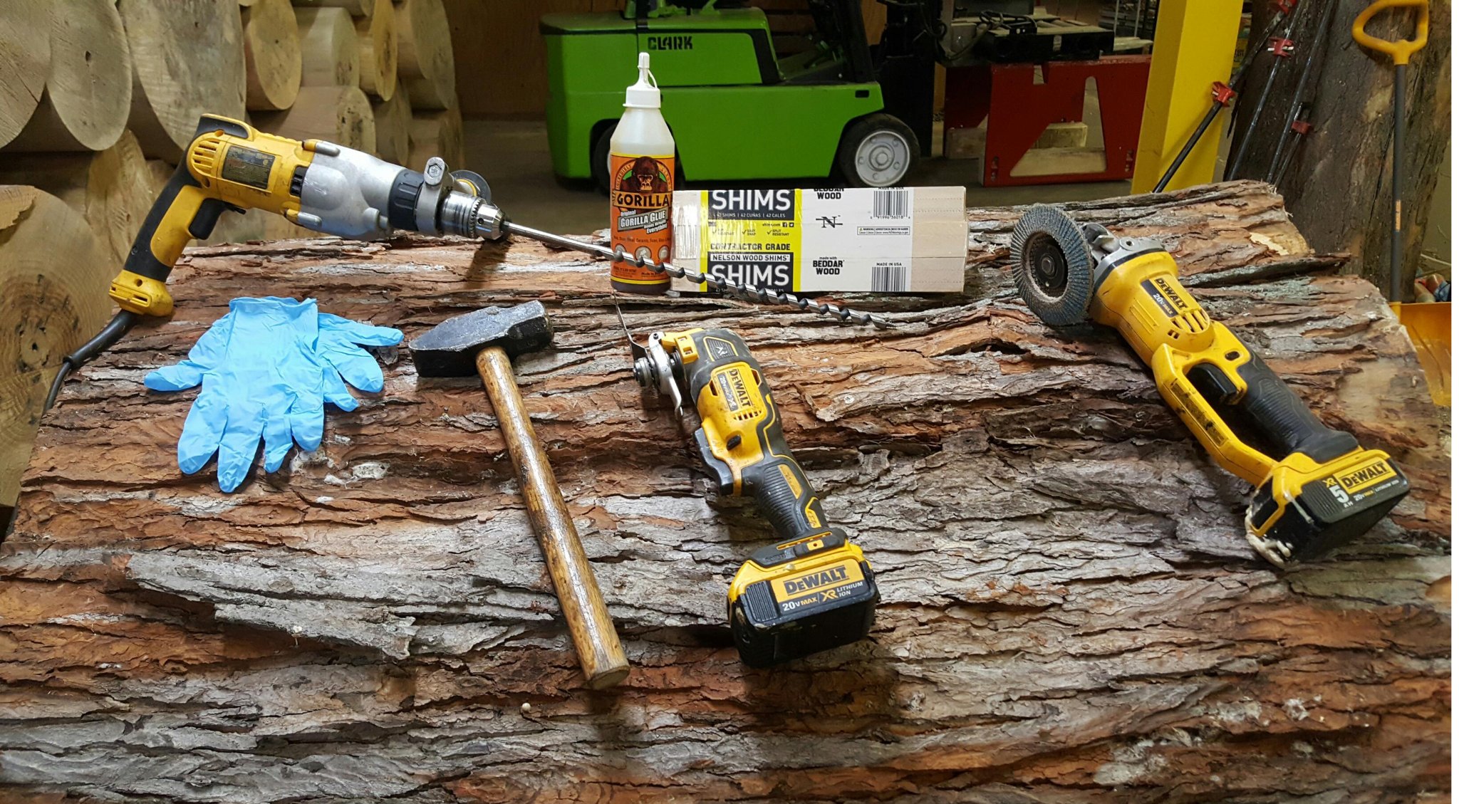 Full IC WOOD crack repair tool kit laid out on a bark-on log: DeWalt hammer drill with long auger bit, DeWalt oscillating multi-tool, DeWalt angle grinder with flap disc, Gorilla Glue, Nelson cedar wood shims, rubber mallet, nitrile gloves — green Clark forklift and wall of finished logs visible in background